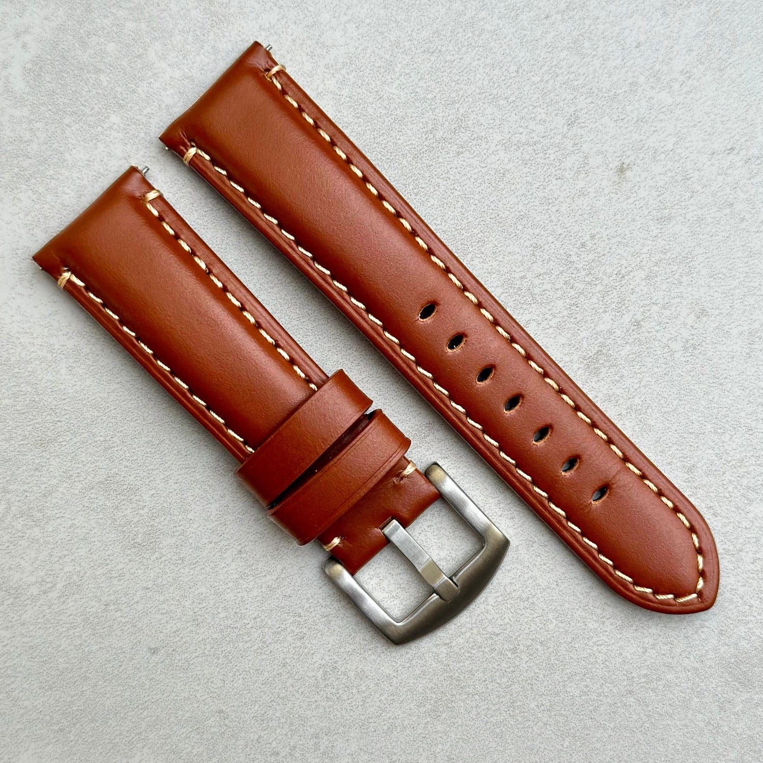 Oslo dark caramel brown full grain leather watch strap placed diagonally on a grey background. Contrast ivory stitching. Brushed 316L stainless steel buckle. 