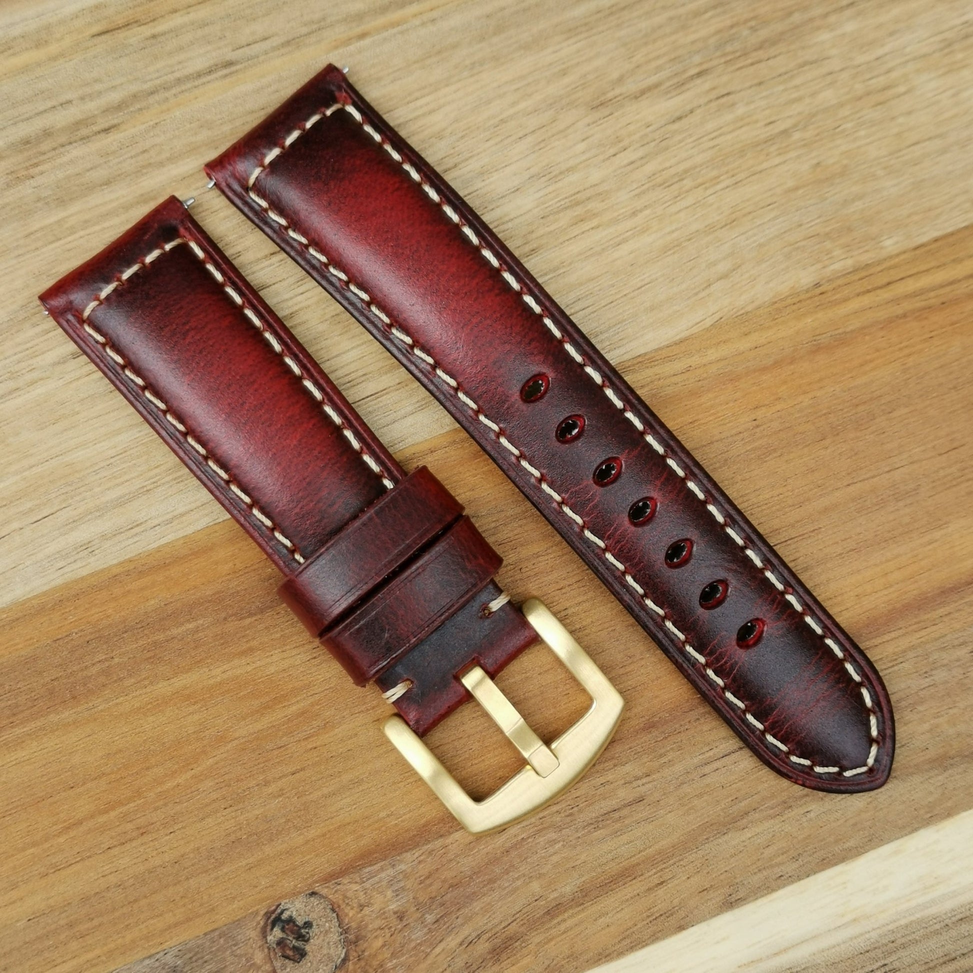 Berlin red padded leather watch strap with gold buckle and contrast ivory stitching. Placed on a wood background.