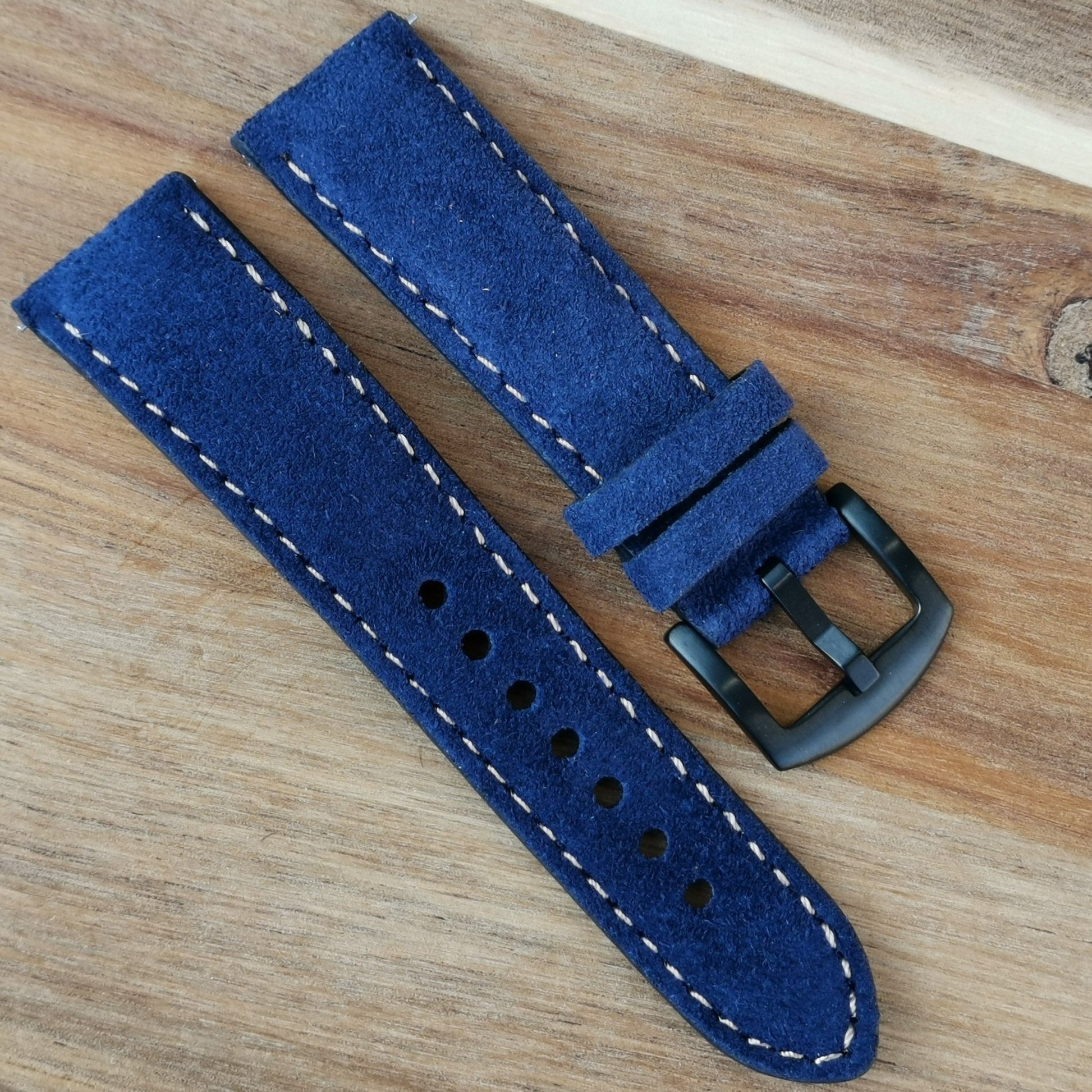 Paris navy blue suede watch strap fitted with an iodized black 316L stainless steel buckle. Placed diagonally on a wood background.